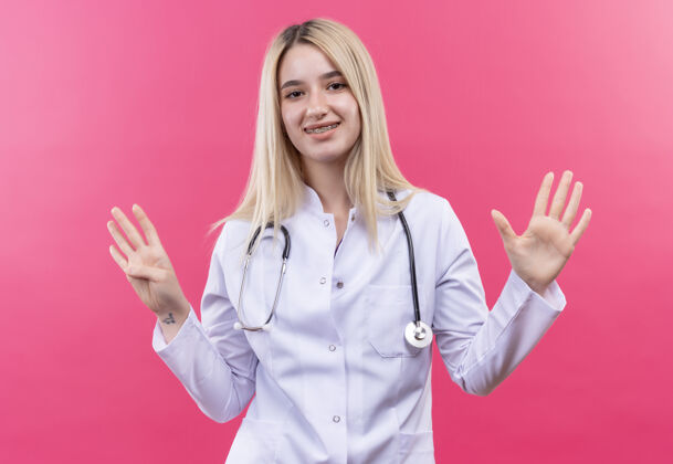 穿着微笑的医生年轻的金发女孩穿着医用长袍戴着听诊器和牙套显示不同的数字 手放在孤立的粉红色背景上手粉红色支架