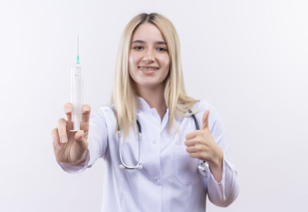 白人微笑的医生年轻的金发女孩戴着听诊器和医用长袍戴着牙套拿着注射器她的拇指在孤立的白色背景上拇指穿着女孩