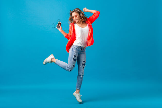 长度迷人的微笑快乐的女人戴着耳机跳舞听音乐穿着时髦风格的服装在蓝色的工作室背景下隔离 穿着粉色夹克和太阳镜跳跃运动年轻