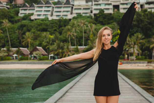 模特穿着黑色连衣裙的美丽迷人的女人在豪华度假酒店的码头上摆姿势 暑假 热带海滩高大放松风