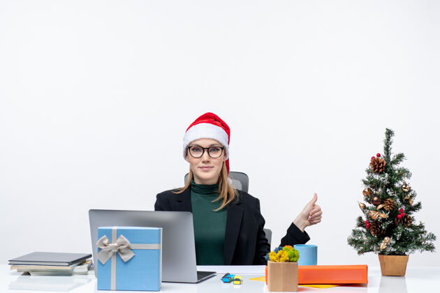 桌子一位微笑的商务女士 戴着圣诞老人帽 坐在一张桌子旁 桌上放着圣诞树和礼物 指着办公室左侧白色背景上的东西礼物坐着一边