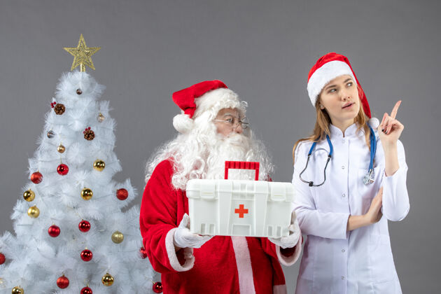 快乐圣诞老人和给他急救箱的女医生在灰色墙上的正视图圣诞老人第一庆祝