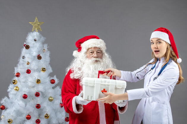 人圣诞老人和给他急救箱的女医生在灰色墙上的正视图女性第一前面