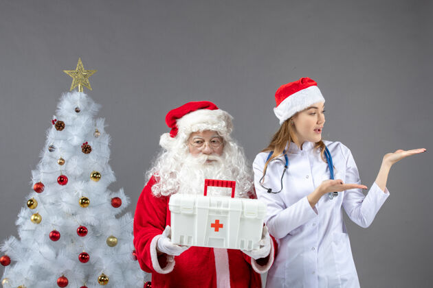 人圣诞老人和给他急救箱的女医生在灰色墙上的正视图人们谁男人
