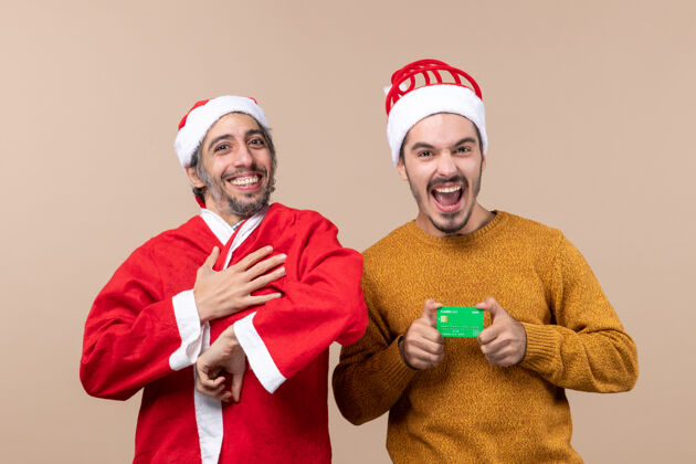 帽子正面图两个快乐的朋友一个穿着圣诞老人的外套 另一个是米色背景的信用卡风景前面男人