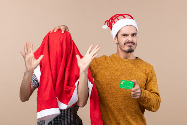 两个正面图两个家伙一个头上套着圣诞老人的外套 另一个拿着一张卡片 在米色孤立的背景上抚摸着他的朋友的头前面米色伙计们