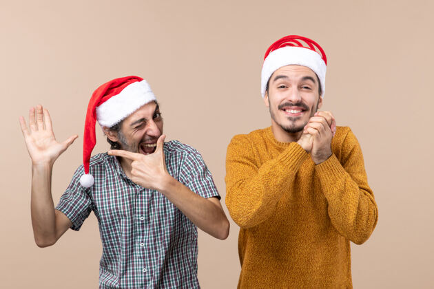 封闭前视图两个戴着圣诞帽的家伙一个展示他的手另一个闭着眼睛许愿米色孤立的背景手展示圣诞老人