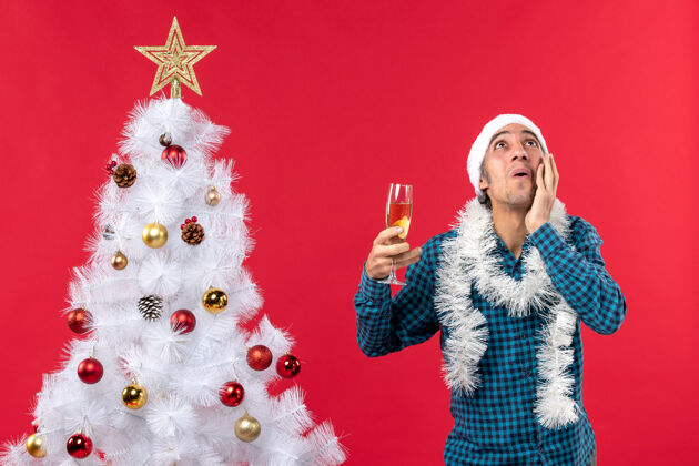 看圣诞节心情与情绪激动的年轻人与圣诞老人帽子在一个蓝色的剥夺衬衫举行了一杯葡萄酒向下看圣诞树附近红色年轻人帽子