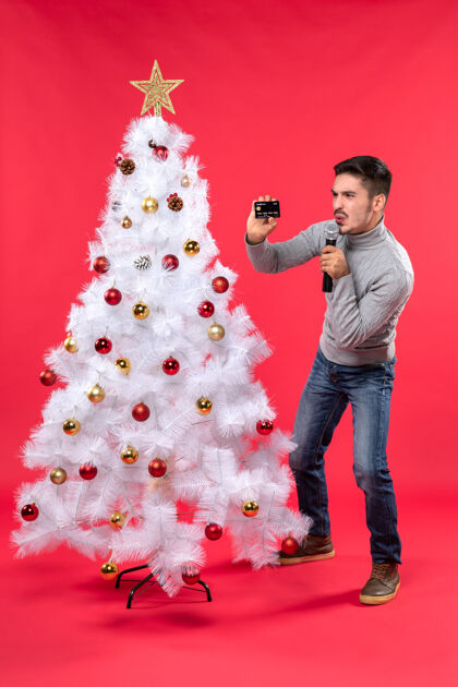 情绪化圣诞节心情与情绪化的家伙站在装饰圣诞树附近 拿着麦克风和电话与搞笑的手势雪人麦克风圣诞