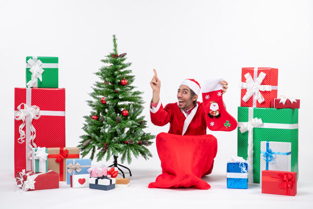 惊讶惊喜的圣诞老人正指着上面坐在地上 拿着圣诞袜靠近礼物和装饰在白色背景上的新年树圣诞老人礼物帽子