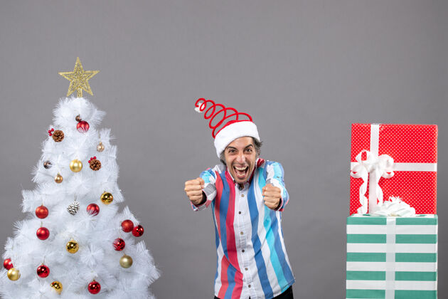 快乐在白色圣诞树旁 戴着螺旋弹簧圣诞帽的年轻人兴高采烈地展示着他的幸福圣诞树圣诞快乐