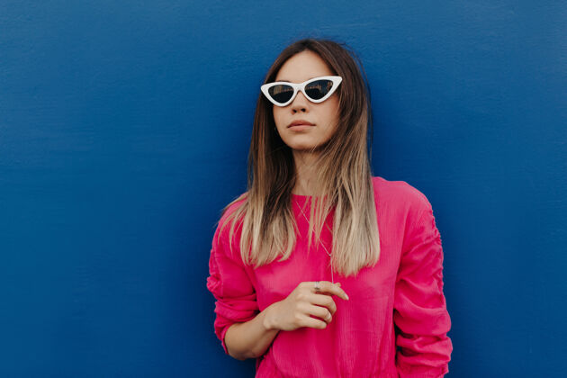 太阳镜特写户外肖像迷人的年轻女子穿着粉红色衬衫和白色太阳镜摆在孤立的蓝色墙壁摆姿势年轻快乐