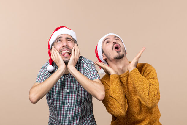 两个正面图两个戴着圣诞帽的吓坏了的家伙一个捂着耳朵 另一个一边大声喊叫一边看着高高的米色孤立背景伙计们掩护圣诞老人
