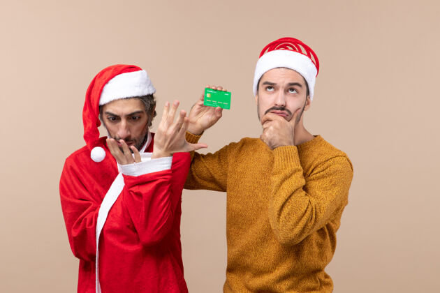 看前视图两个朋友一个穿着圣诞外套看着他的手 另一个拿着卡在米色孤立的背景上朋友两个男性