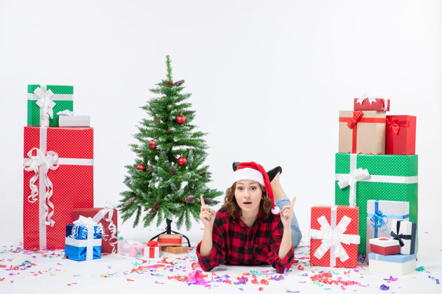 庆祝年轻女子躺在白色墙壁上的圣诞礼物和小圣诞树周围的正视图前面圣诞快乐铺设