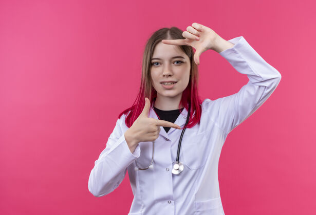 手势穿着听诊器医用长袍的年轻医生女孩很高兴 在粉色孤立的背景上显示出框架姿态女孩听诊器镜框