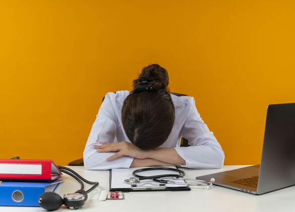 女年轻的女医生穿着医用长袍 手持听诊器 坐在办公桌旁 拿着医疗工具在电脑上工作 低下头 隔离黄色背景长袍医疗医生