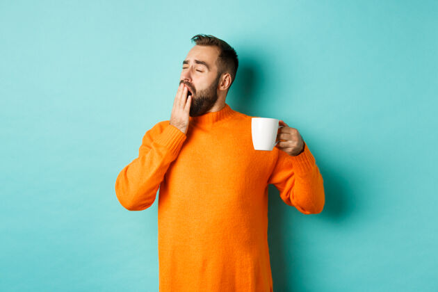 胡须昏昏欲睡的帅哥喝着咖啡打着哈欠 穿着橘色毛衣站在浅蓝绿色的墙上胡须英俊饮料
