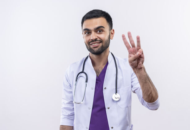 微笑微笑着的年轻男医生穿着听诊器医用长袍 在孤立的白色背景上显示三个听诊器长袍展示
