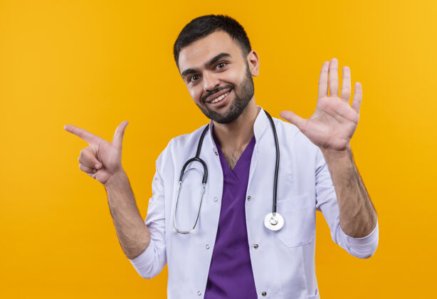 侧面微笑着的年轻男医生穿着听诊器医用长袍 指着旁边 在孤立的黄色背景上显示停止手势医生展示长袍