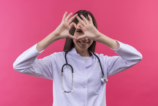微笑微笑的年轻医生女孩穿着听诊器医用长袍 在孤立的粉红色背景上显示出心脏姿态心脏听诊器年轻