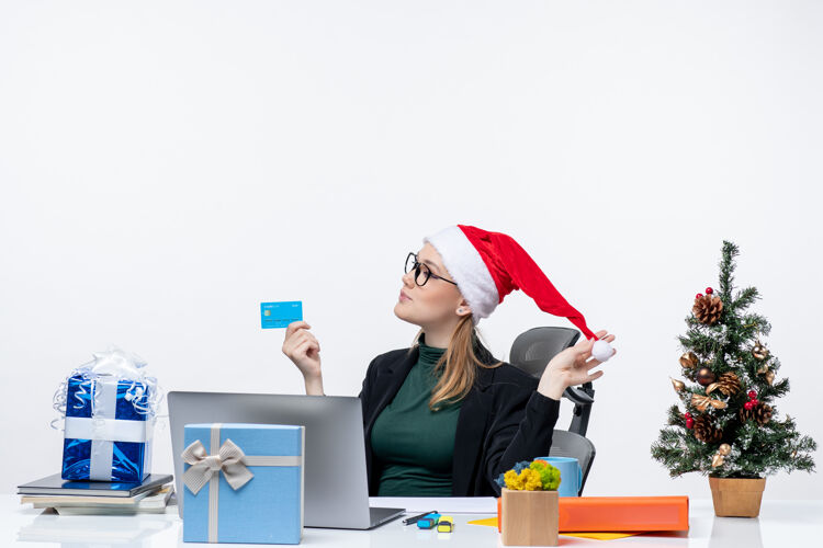 眼镜自信迷人的女人 戴着圣诞老人的帽子 戴着眼镜坐在桌子旁 手里拿着银行卡在办公室里抱着帽子女人