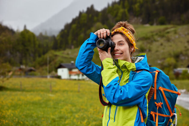 嬉皮士微笑的女人漫步在美丽的山乡 把相机放在眼前 穿着休闲夹克拍照背包冒险摄影