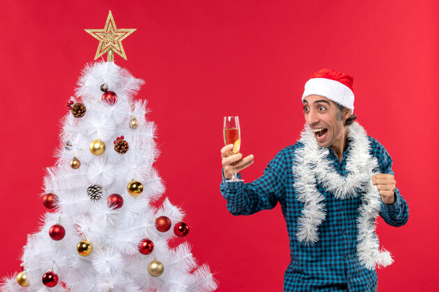 圣诞老人圣诞节心情与疯狂的情绪年轻人圣诞老人帽子在一个蓝色条纹衬衫举行了一个靠近圣诞树的葡萄酒杯人年年轻人