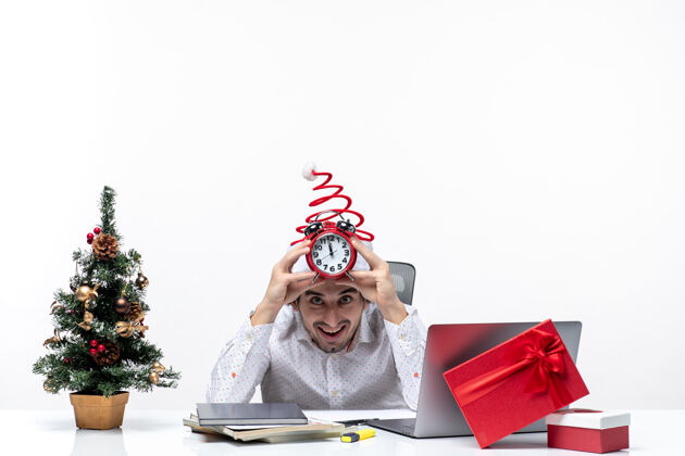 办公室戴着圣诞老人帽 拿着闹钟的年轻商人惊奇地看着什么东西 坐在白色背景的办公室里惊喜礼物时钟