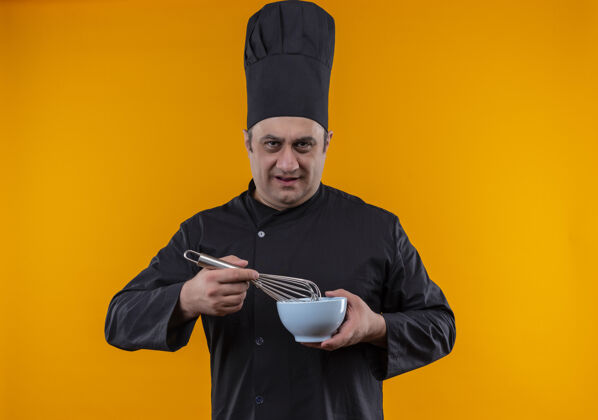 厨师严格的中年男性厨师 身着厨师制服 手持黄色背景的搅拌器和碗严格碗拂