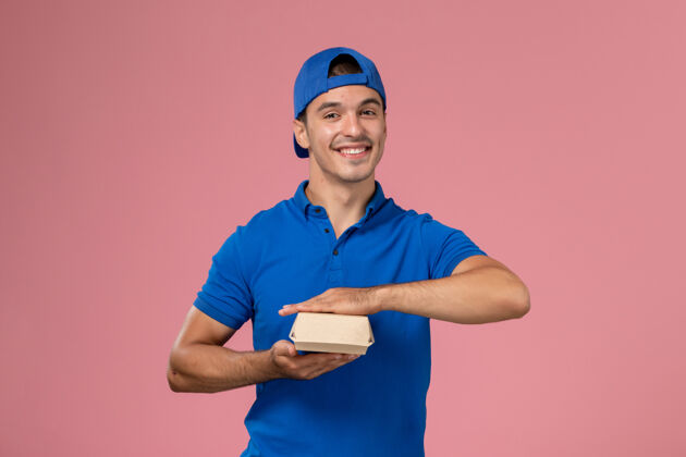 送货正面图身穿蓝色制服披肩的年轻男性快递员拿着小快递食品包微笑着站在粉红色的墙上食物快乐服务