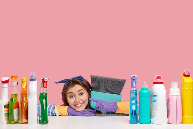 清洁剂满意的女佣横拍戴着头巾 指着洗涤剂 紧握着扫帚 喜欢它的完美效果牙齿清洁地板