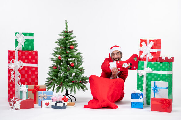 地有趣的圣诞老人坐在地上 穿着圣诞袜 靠近礼物和装饰在白色背景上的新年树圣诞老人礼物季节性