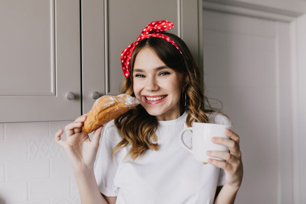 微笑漂亮女孩吃甜羊角面包的室内照片迷人的欧洲女士在午餐时玩得很开心的照片可爱舒适年轻