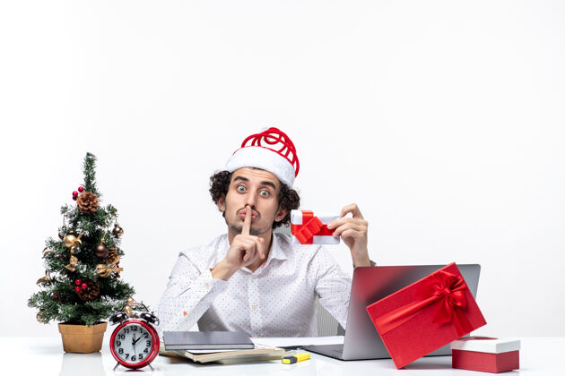 手势圣诞节心情惊讶的商人与圣诞老人的帽子 提高他的礼物 并在白色背景沉默的姿态商务帽子微笑