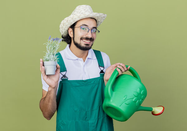 穿着年轻的留着胡须的园丁 穿着连体衣 戴着帽子 手里拿着浇水罐 手里拿着盆栽植物 满脸笑容帽子园丁光