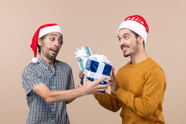两个正面图两个戴着圣诞帽的快乐的家伙在米色孤立的背景上交换礼物帽子圣诞老人前面