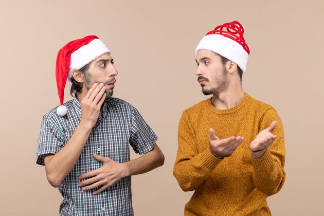 前面正面图两个戴着圣诞帽的困惑的家伙在米色孤立的背景下看着对方视图男人困惑