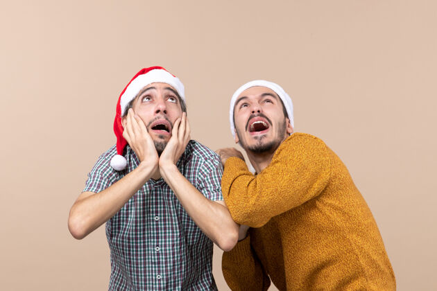米色正面图两个戴着圣诞帽的吓坏了的家伙一个捂着耳朵看着高高的米色背景伙计们害怕帽子