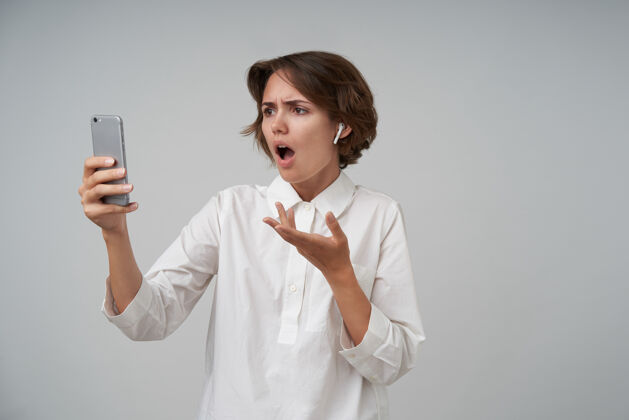 女人年轻忧郁的女性的肖像 随意的发型 举着手机 打视频电话 看着屏幕 一脸震惊 穿着正式的衣服理发摆姿势正式