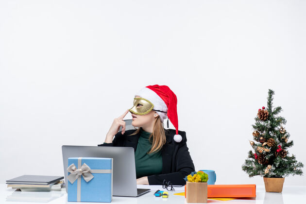 坐着带着圣诞老人帽戴着面具的体贴的年轻女子坐在白色背景的桌子旁 感受圣诞节的气氛桌子圣诞老人圣诞老人