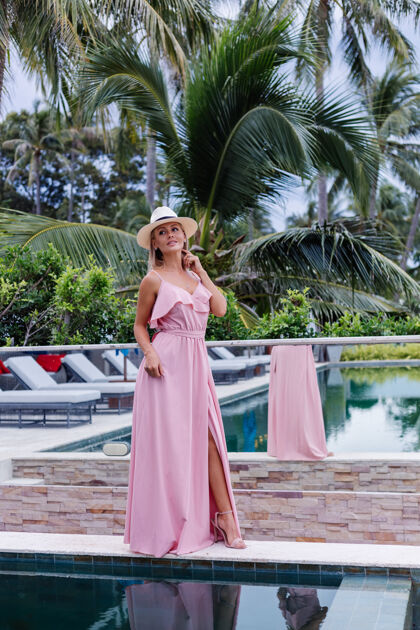 海洋穿着浪漫优雅粉色长裙的白种女人的肖像 在豪华的别墅酒店度假 美丽的热带棕榈树映衬着戴着经典白帽子的女人优雅露台热带