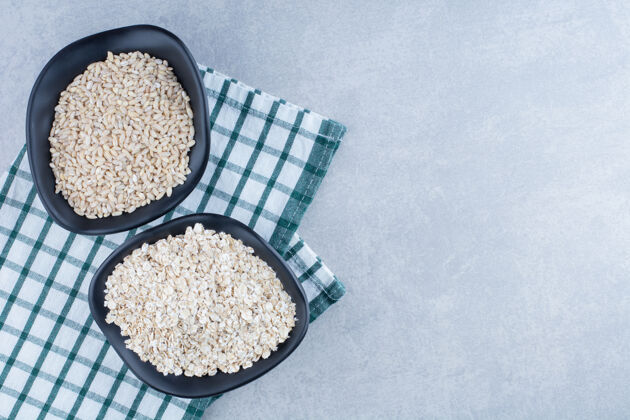 米饭短粒米饭和燕麦堆在两个黑色的碗里 放在大理石背景的折叠桌布上燕麦烹饪桌布
