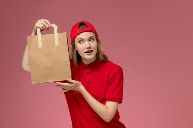 年轻女快递正面图：身穿红色制服 披风的年轻女快递员在粉色墙上拿着快递食品包女人包裹帽子