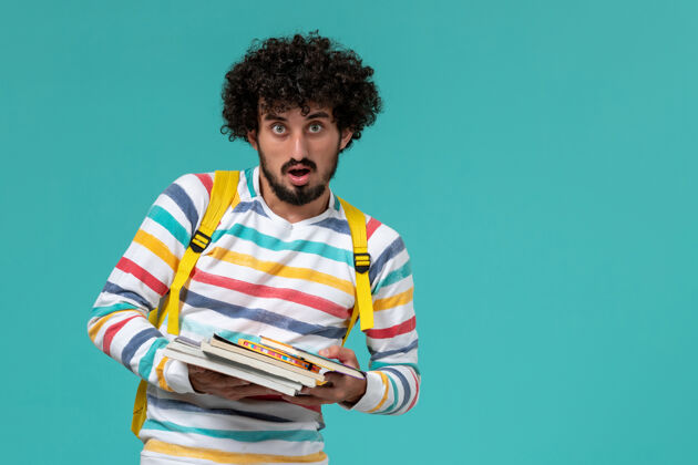 学校身穿条纹衬衫 背着黄色背包 在蓝色墙上拿着书的男学生的正视图画像学生人
