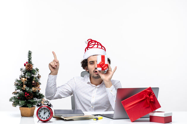 孩子圣诞节心情与有趣的商人与圣诞老人的帽子举起他的礼物 他的脸 并指出以上白色背景圣诞老人圣诞节人