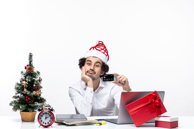 商务带着圣诞老人帽子 留着胡子的年轻商人在办公室里拿着银行卡 把手放在下巴下面 节日的喜庆气氛人年轻生活