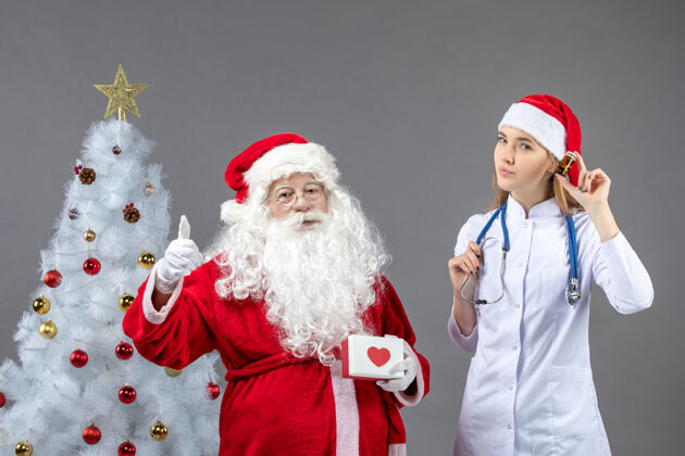 举行圣诞老人和女医生的前视图 女医生拿着装着药片的小瓶子在灰色的墙上女前面服装