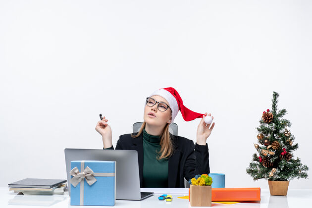 圣诞树新年的气氛与梦幻金发女人坐在一张桌子上 圣诞树和一个白色背景的礼物圣诞老人帽子坐着女人秘书