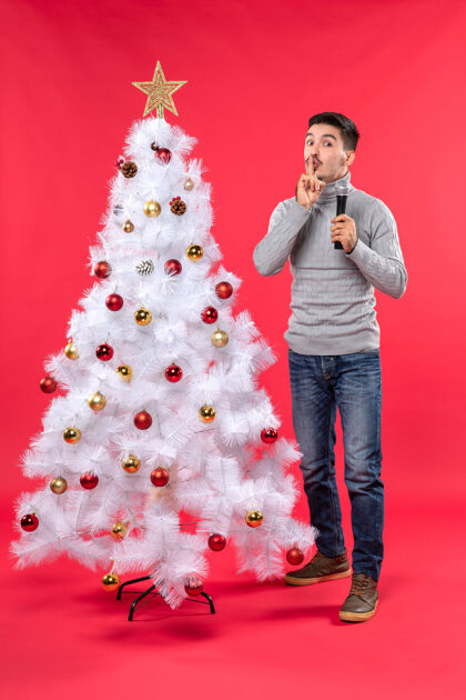 礼物圣诞气氛积极的家伙穿着牛仔裤站在装饰圣诞树附近 拿着麦克风 做着沉默的手势沉默手势制作